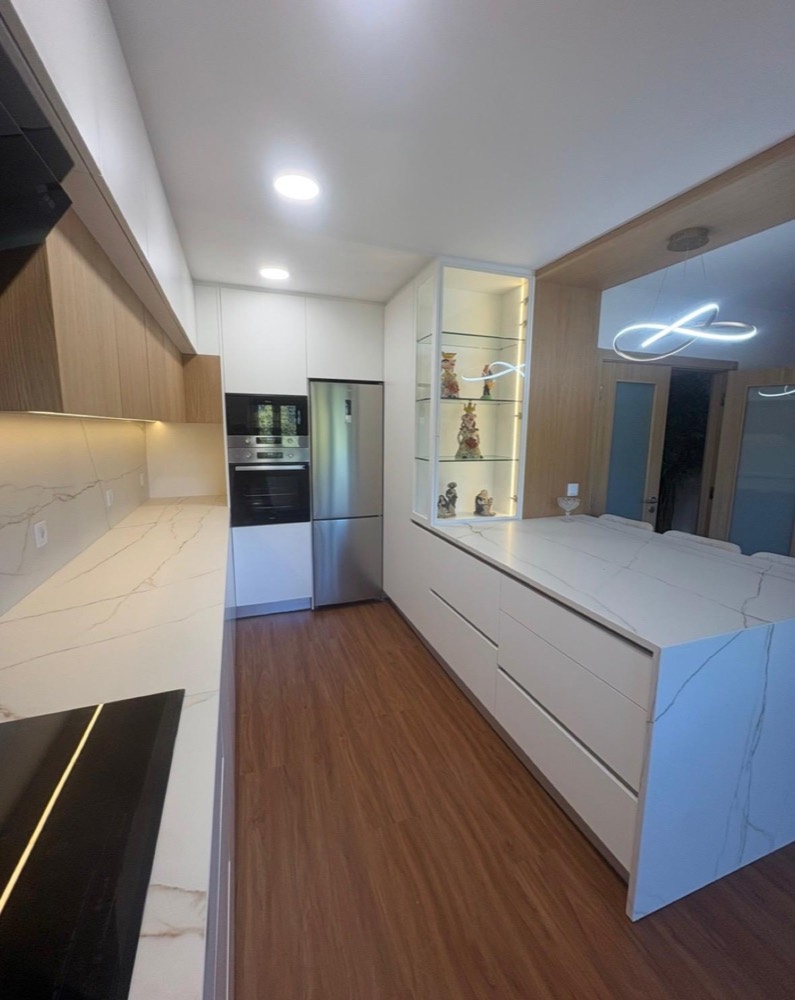 Vista ao longo da cozinha: bancada, colunas de forno e frigorífico, ilha e pavimento em madeira