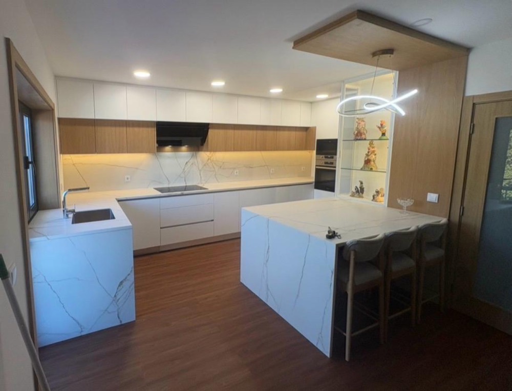 Cozinha remodelada com móveis brancos e em madeira, bancada e ilha em pedra branca com veios e iluminação embutida